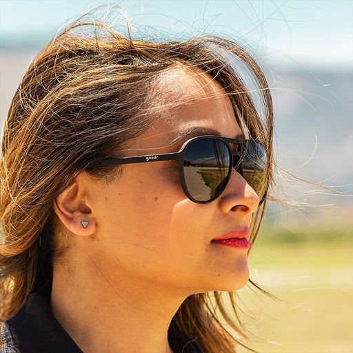 Una donna con una tuta nera e occhiali da sole neri da aviatore con lenti nere non riflettenti guarda avanti con fierezza.