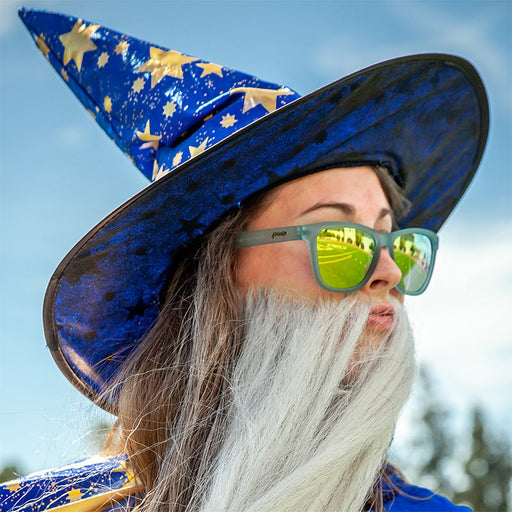 Een vrouw met een tovenaarshoed en grote nepbaard kijkt naar buiten, ze draagt een doorschijnende lichtblauwe zonnebril met gouden reflecterende glazen.
