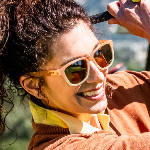Een vrouw met een bruin-witte gradiëntzonnebril zwaait met een golfclub en glimlacht in een zonnestraal.