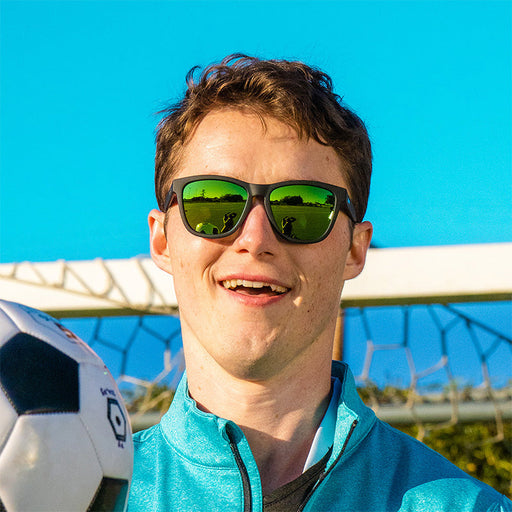 Ein lachender Mann, der eine schwarze Sonnenbrille mit grünen, reflektierenden Gläsern trägt, blickt nach vorne und hält einen Fußball vor ein Tor.