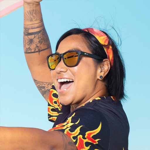 Een vrouw met een zwarte zonnebril met amberkleurige reflecterende glazen, een shirt met vlammotief en een hoofdband lacht.